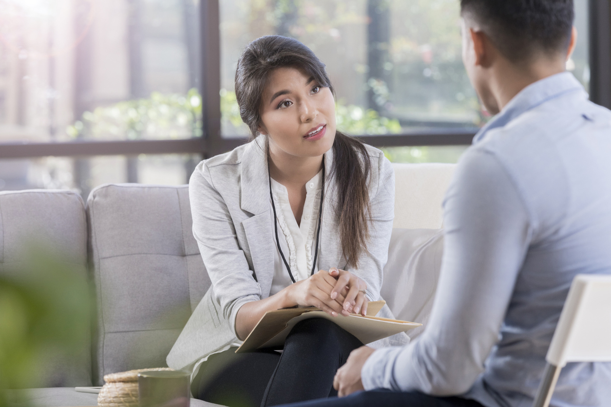 While sitting in a comfortable waiting room, female doctor listens to her patient about mental health problems. Doctor is on a sofa, wearing a lanyard and holding a clipboard.